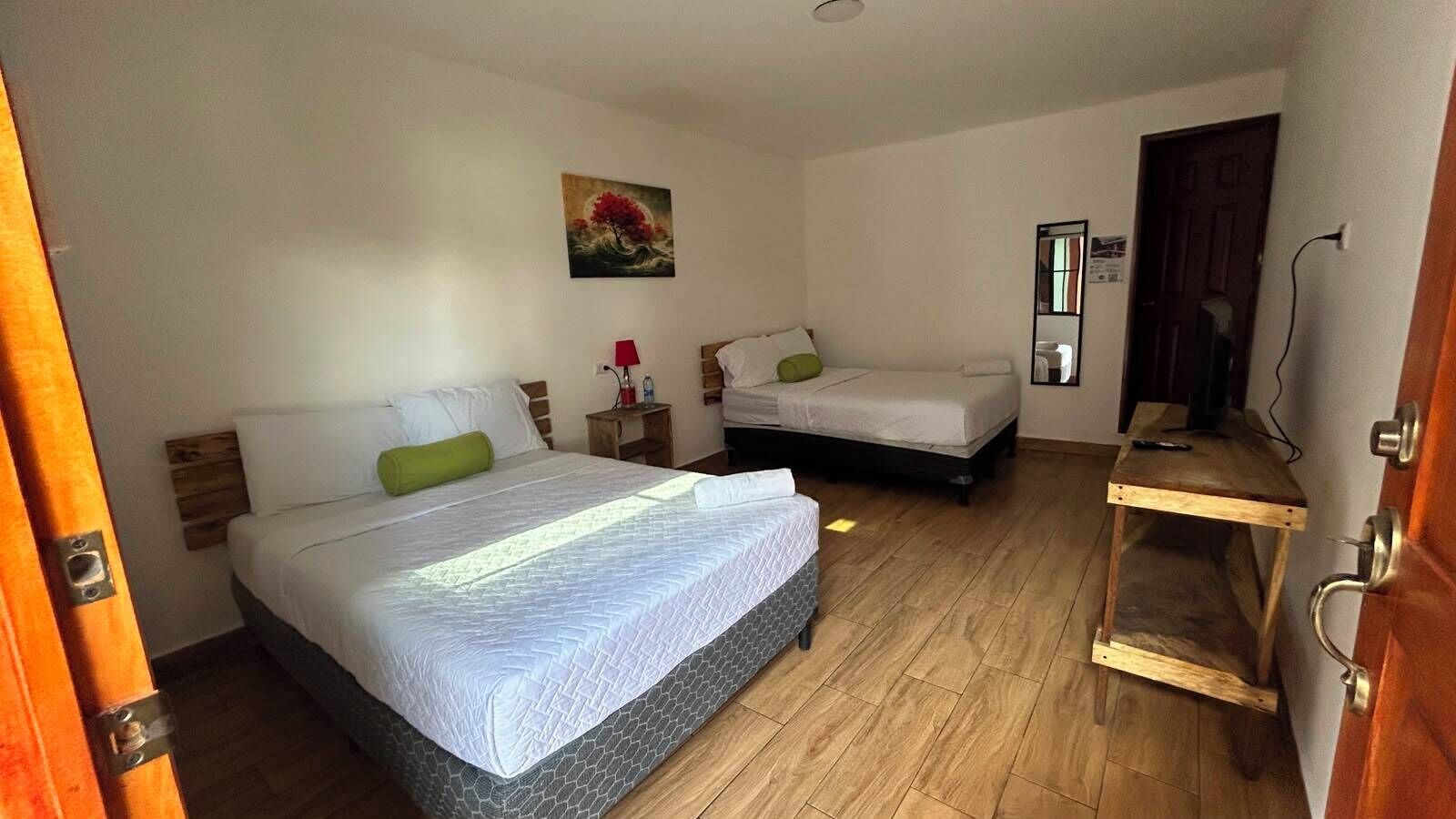 Una habitación con dos camas, piso de madera, un cuadro en la pared y un pequeño estante de madera.
