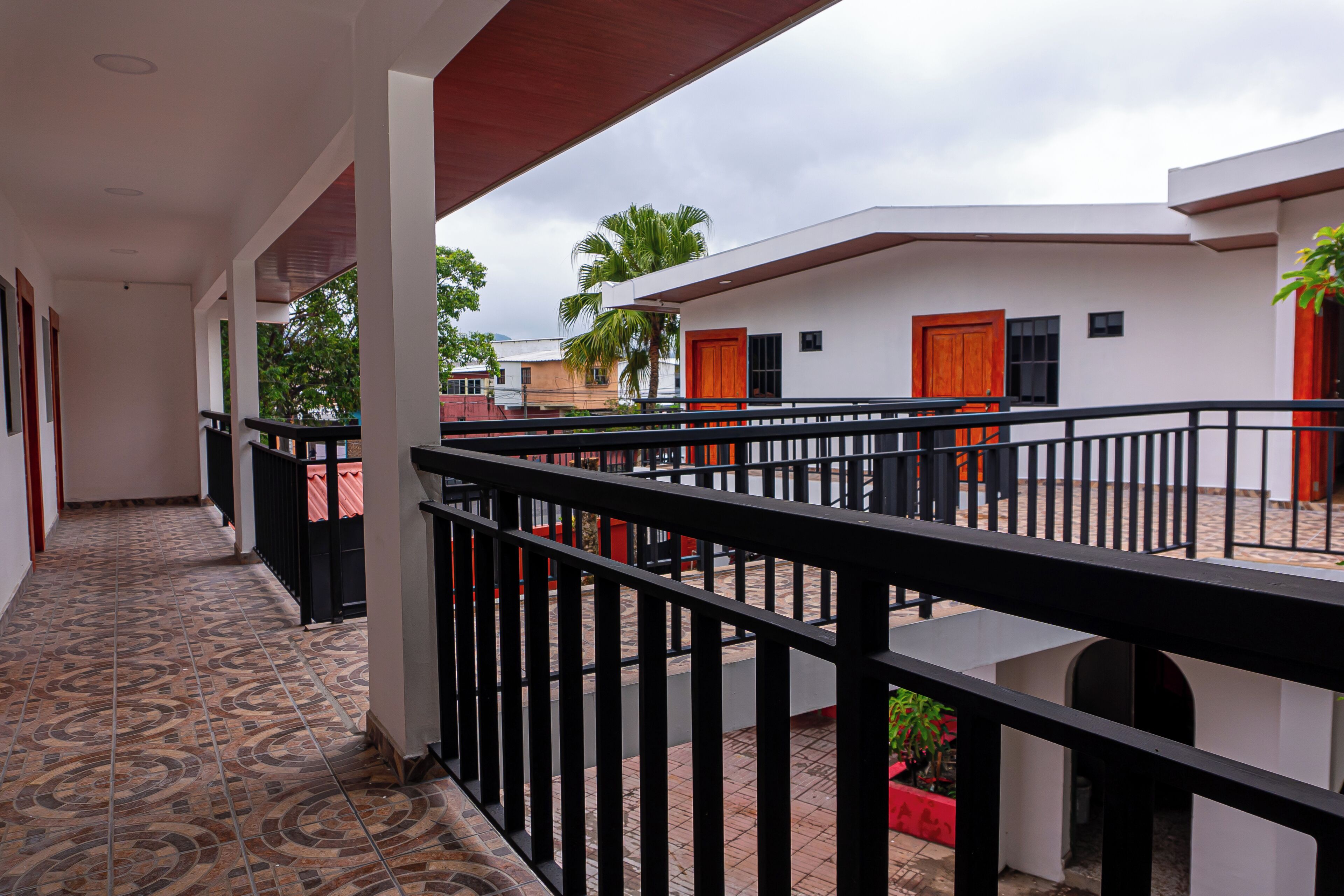 A covered outdoor area with a tiled floor, a black railing, and buildings with red doors in the background.