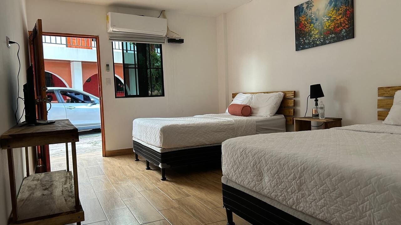 Una habitación de hotel con dos camas, un televisor sobre un mueble, una ventana con vistas a un coche y un edificio, y un cuadro en la pared.