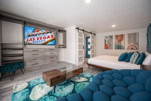 Un dormitorio moderno con cama, televisor mostrando 'WELCOME TO Fabulous LAS VEGAS', cómoda, banco y arte abstracto en la pared.