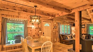 Interior de una cabaña de madera con comedor, sala de estar con chimenea y ventanas amplias con cortinas a cuadros.