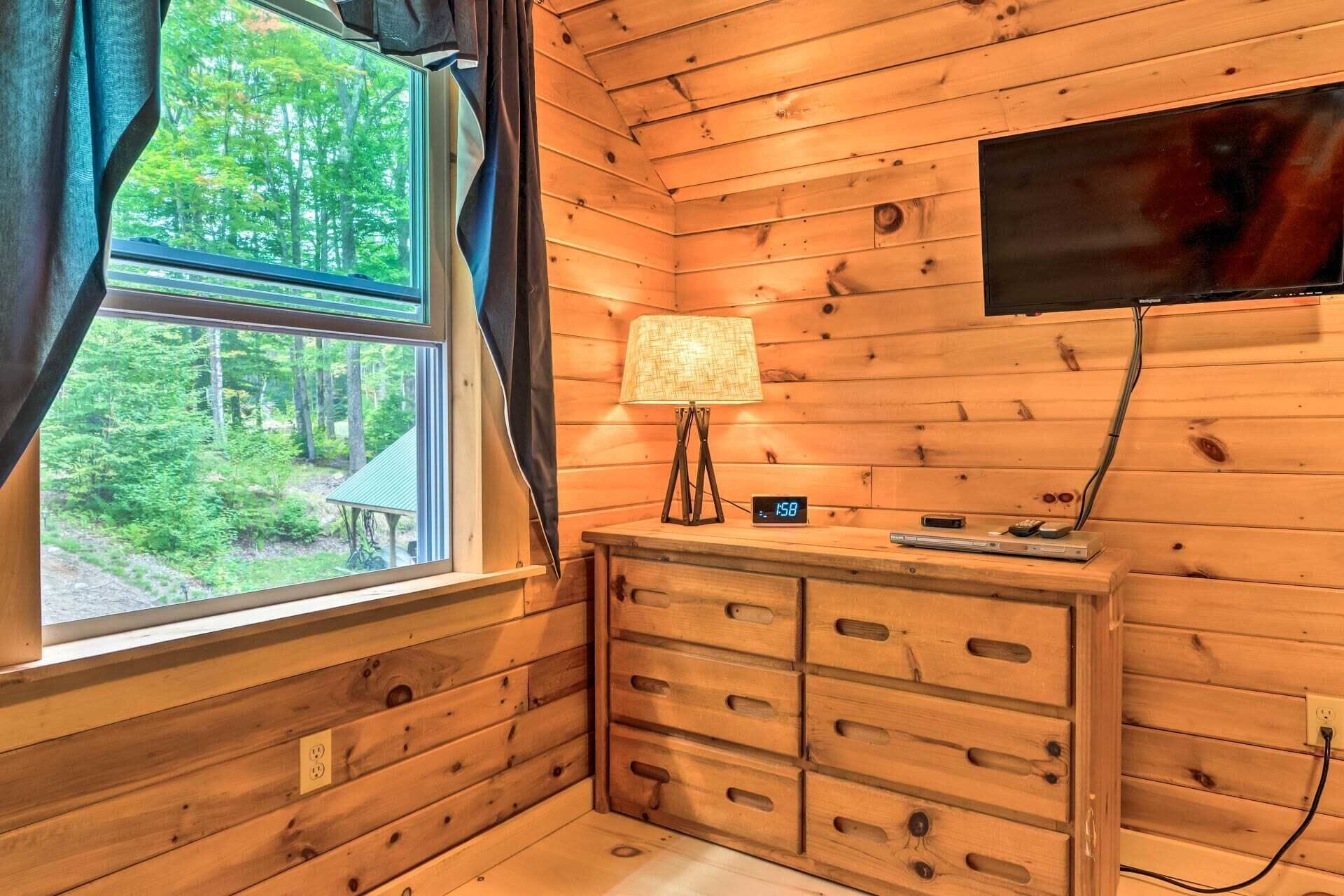 Interior de uma cabana com janela, luminária, relógio e TV de tela plana na parede.