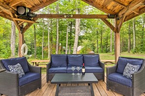 A covered outdoor seating area with wicker furniture and a table, surrounded by a forest.