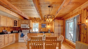 Cocina de cabaña de madera con mesa de comedor, sillas, microondas, estufa y un ventanal grande con cortinas.