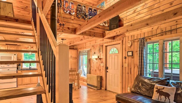Interior de una cabaña de madera con escalera, electrodomésticos de cocina, chimenea y un sofá de cuero.