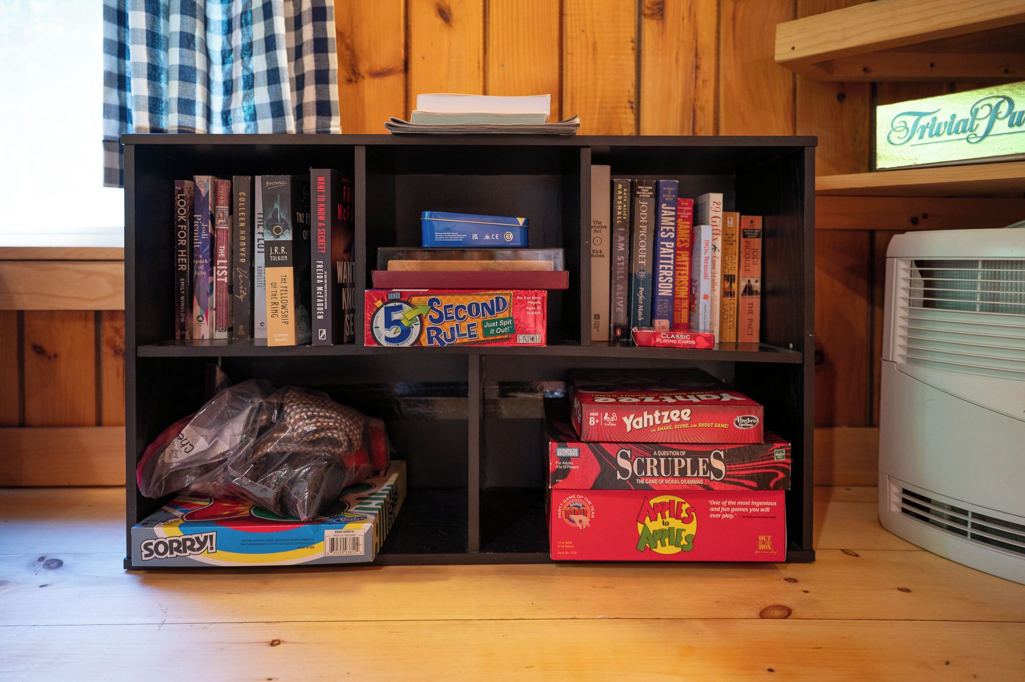 Una estantería con varios libros y juegos, una bolsa de bocadillos y un pequeño aire acondicionado.