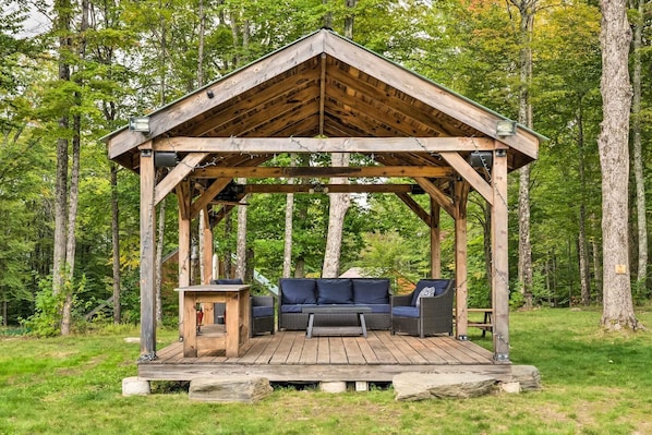 A wooden gazebo with a bench and table, surrounded by a forest.