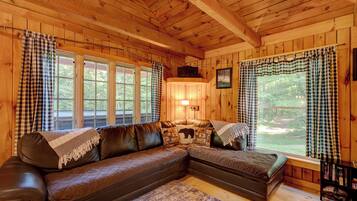 A cozy cabin interior with a large L-shaped leather sofa, a fireplace, and a large window with checkered curtains.