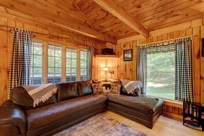 Interior acogedor de una cabaña con un amplio sofá de cuero en forma de L, una chimenea y un ventanal con cortinas de cuadros.