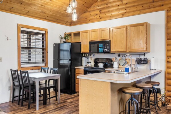 A compact kitchen with wooden cabinets, a black refrigerator, a microwave, and a stove. There is a dining table with chairs and a small bar area with stools.