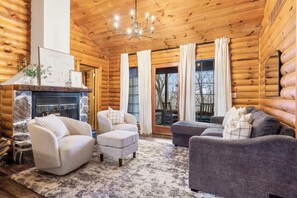 A log cabin interior with a fireplace, two sofas, a rug, and a chandelier.