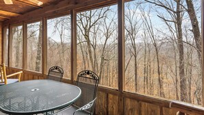 Una habitación con paneles de madera, una mesa redonda de metal y dos sillas, con amplios ventanales que dan a un bosque.