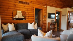 Un acogedor interior de cabaña de madera con un sofá seccional gris, un sillón blanco y techo de madera.
