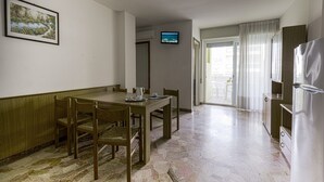 A dining area with a wooden table and chairs, a television on the wall, and a kitchen area with a refrigerator and cabinets.
