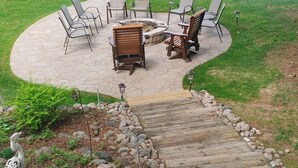 Een rond zitgedeelte buiten met stoelen en een vuurkorf, omgeven door een stenen terras en een houten vlonder die naar een zwembad leidt.