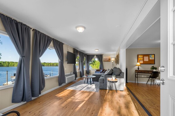 A spacious living area with a large window overlooking a body of water, a sofa, a coffee table, and a desk with a lamp.