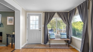 A room with a wooden floor, a bench with pillows, a door, and large windows with curtains.