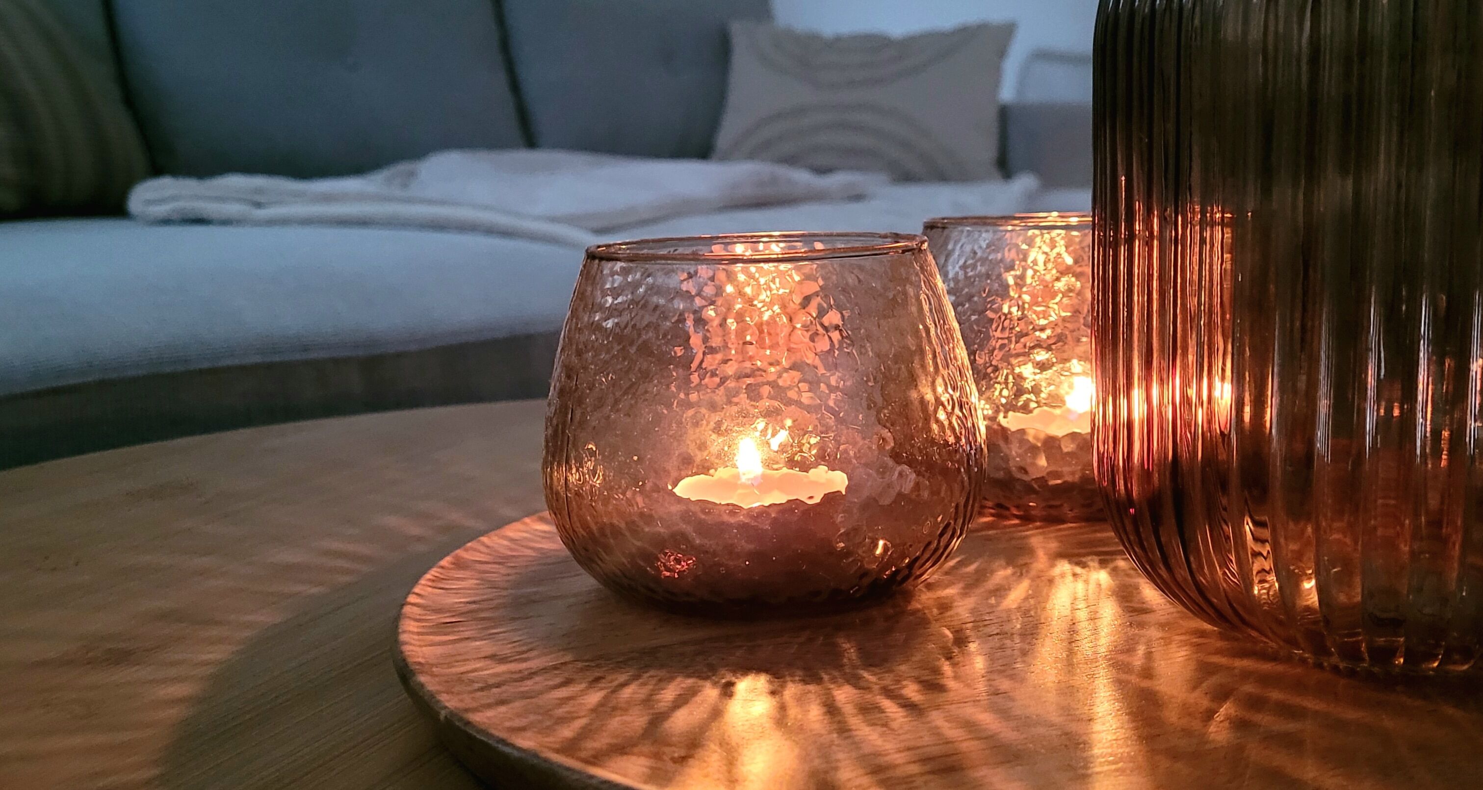 A candlelit table with two candles in glass holders, a wooden coaster, and a sofa in the background.