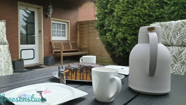 A table set with a slice of chocolate cake, a white mug, and a white teapot on a wooden table outdoors. - Luette Ostfreesenstuuv (Holtgast)