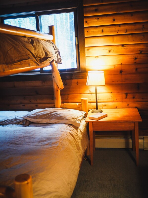 Une chambre chaleureuse dans un chalet, avec un lit superposé, une table de chevet avec une lampe et une fenêtre donnant sur les arbres.