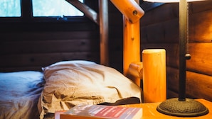 En stuga med träpanel på insidan, med en säng, en lampa och en bok på ett bord.