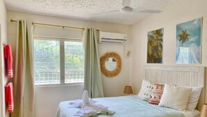 Een slaapkamer met een bed, een raam met gordijnen, een wandairconditioner, een spiegel en twee kunstwerken aan de muur.