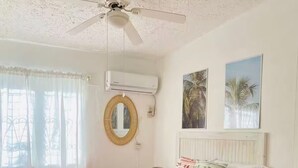 Un dormitorio con cama, aire acondicionado, ventilador de techo, ventana con cortinas y dos cuadros en la pared.