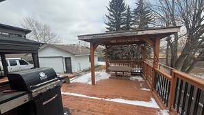 A wooden deck with a grill and a gas tank, a covered seating area, and a view of a house and trees in the background. - Cozy Calgary Stampede Sanctuary Home (Calgary)