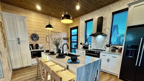 A modern kitchen with wooden cabinets, a marble island, bar stools, pendant lights, and a black refrigerator. - Mountain High Rooftop Cabin Pigeon Forge NEW Indoor Pool Hot Tub Sleeps 15 (Pigeon Forge)