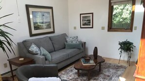 A living room with a grey sofa, a black armchair, a coffee table, and a rug. There are two framed pictures on the wall, a potted plant, and a window with a view of trees. - Seapowet suite (Tiverton)