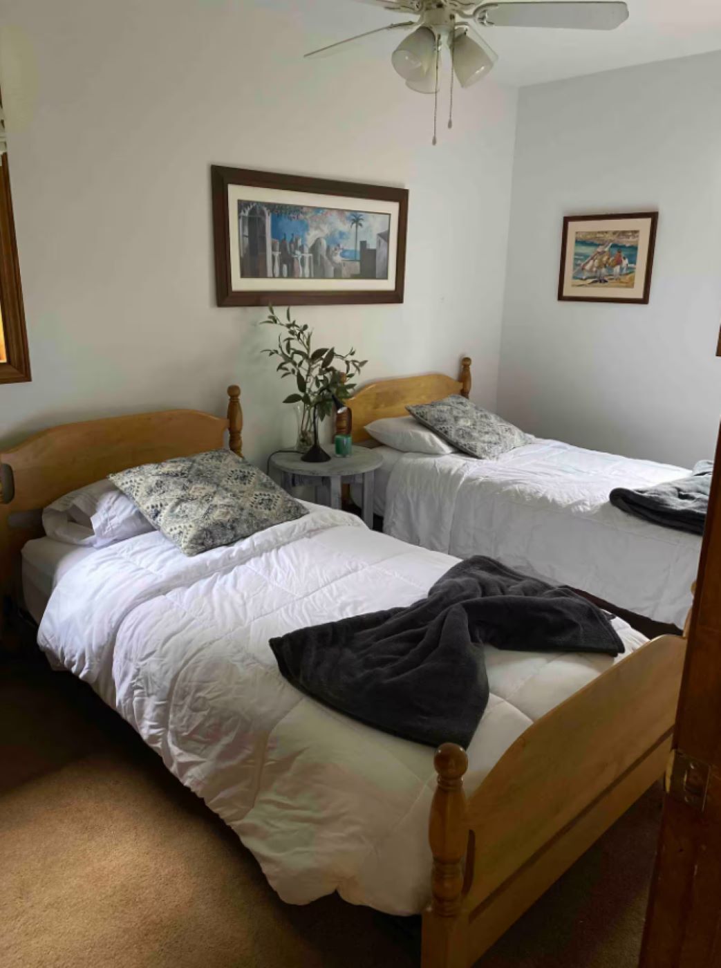 Een kamer met twee eenpersoonsbedden, een plafondventilator, een klein rond tafeltje met een plant en twee ingelijste foto's aan de muur.