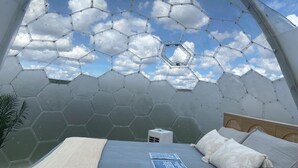 A room with a hexagonal glass ceiling, a bed with a blue bedspread, and a small bedside table with a lamp. - Meadowlark Sky Domes - Sleeps 6 + Private Hot Tub +Cold Plunge Pool! (Cardston)