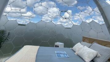 A room with a hexagonal glass ceiling, a bed with a blue bedspread, and a small bedside table with a lamp.