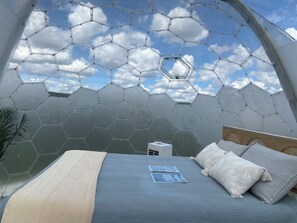 Quarto com teto de vidro hexagonal, cama com colcha azul e uma pequena mesa de cabeceira com abajur.