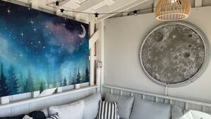 A cozy seating area with a starry night mural, a round moon-like wall decoration, and a wooden pendant light. - Meadowlark Sky Domes - Sleeps 6 + Private Hot Tub +Cold Plunge Pool! (Cardston)