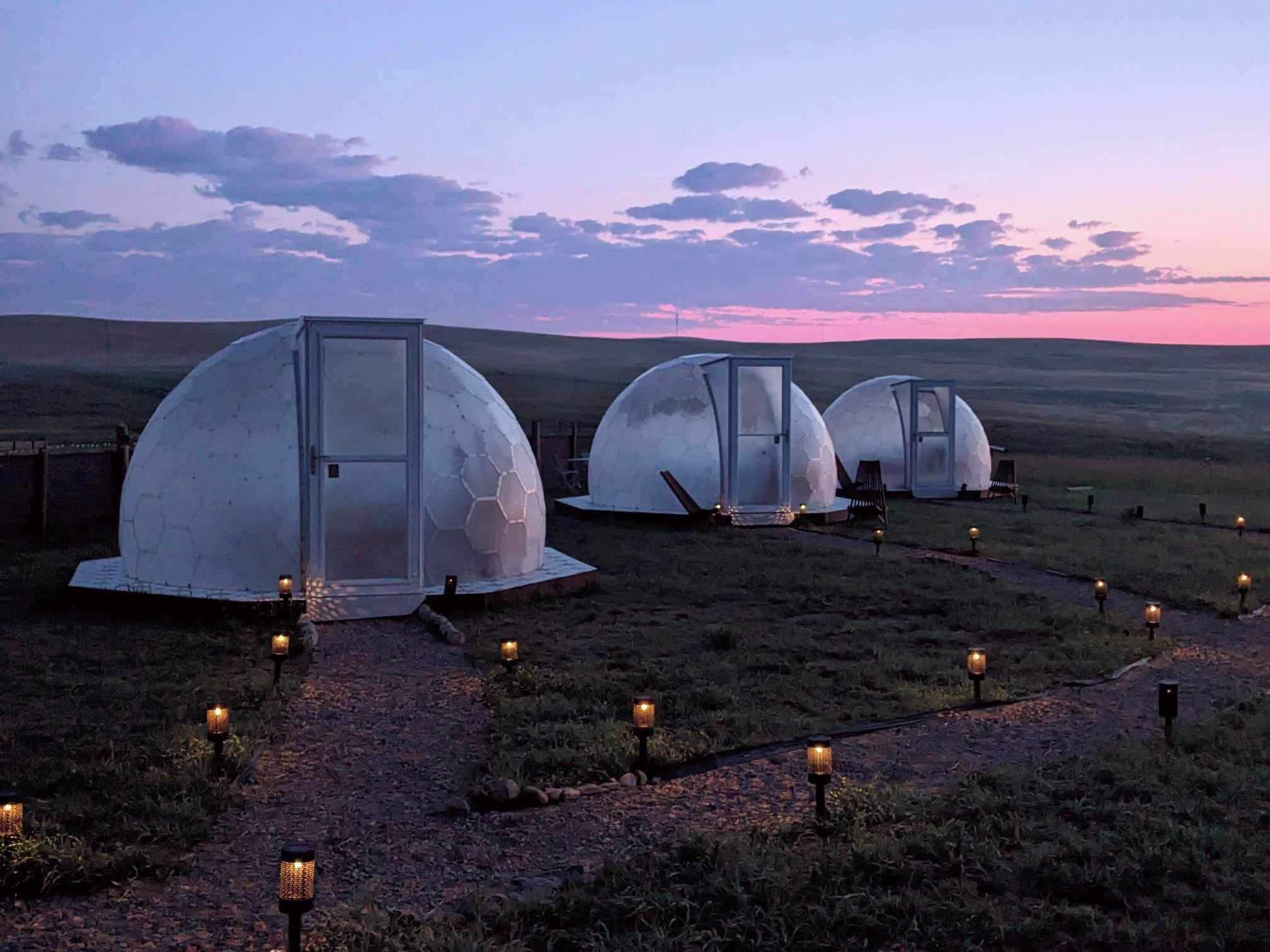 Three dome-shaped structures with glass doors, connected by a gravel path with lanterns, set against a twilight sky.