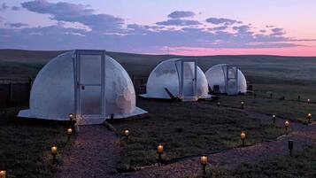 Three dome-shaped structures with glass doors, connected by a gravel path with lanterns, set against a twilight sky.