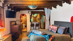 A cozy bedroom with a bed, a desk, a chair, a TV, and a bathroom visible through an archway. - Cozy Private Adobe Studio • Kiva Fireplace • Walk to Taos Plaza • Pet-Friendly (Taos)