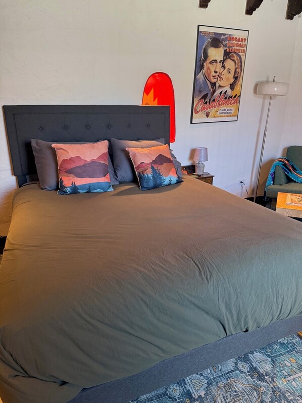 A neatly made bed with a grey bedspread and two decorative pillows, a bedside table with a lamp, and a poster on the wall. - Cozy Private Adobe Studio • Kiva Fireplace • Walk to Taos Plaza • Pet-Friendly (Taos)