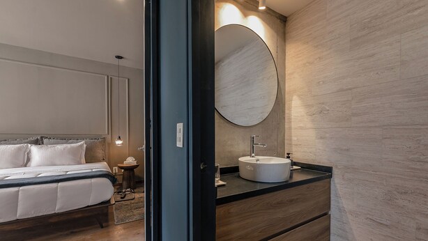 A modern bedroom with a bed, bedside table, and a round mirror above a wooden vanity with a sink. - Indigo Veracruz Roma norte Apt 1B I 1B (Mexico City)