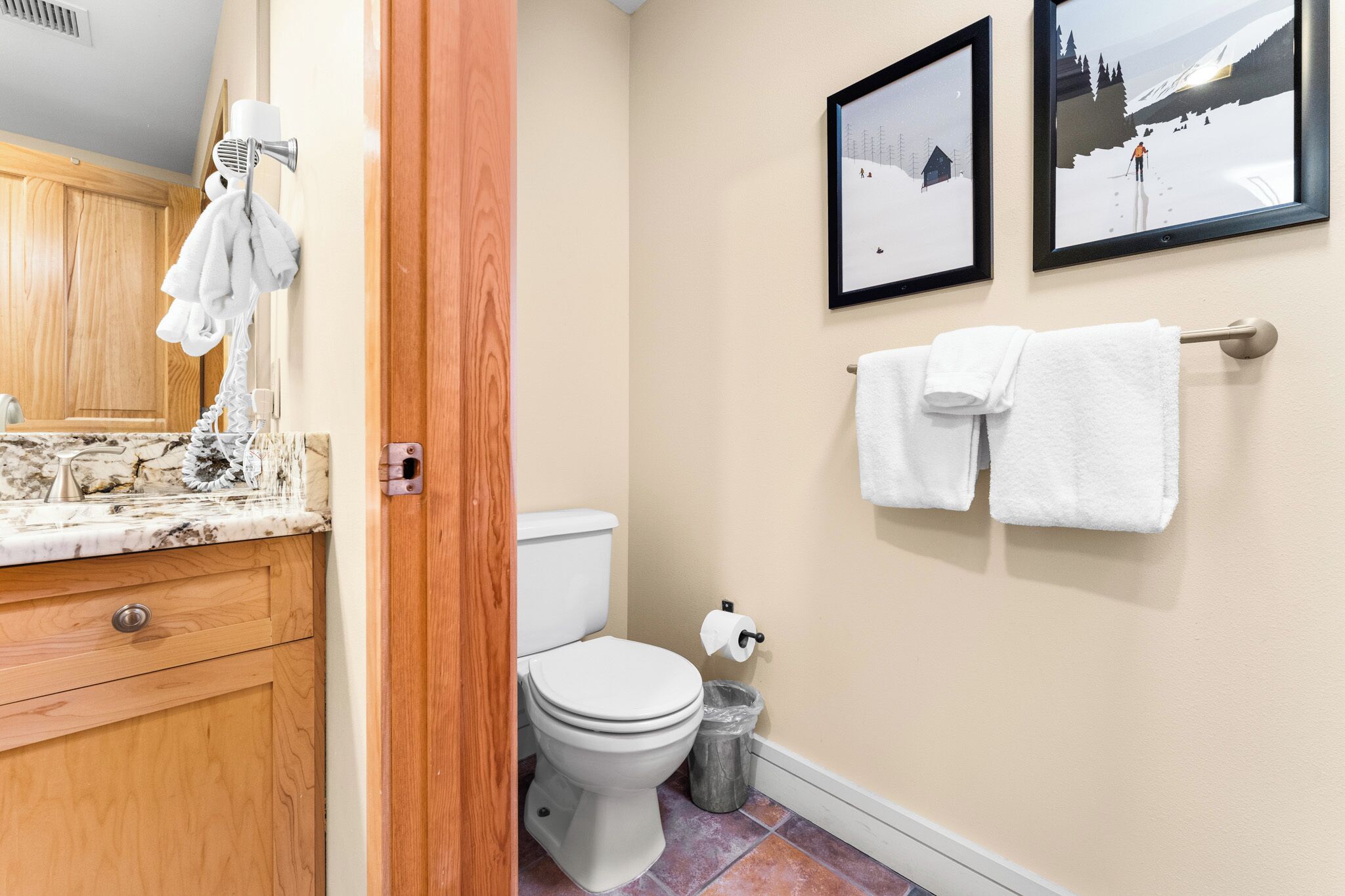 A bathroom with a toilet, a sink with a marble countertop, wooden cabinets, and two framed pictures on the wall.