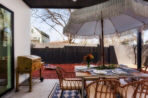 An outdoor dining area with a table set for two, a wicker chair, a small cart, and a large striped umbrella. - Modern East Austin Hideaway - Stylish - Walkable (Austin)