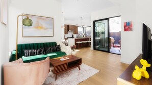 A modern living room with a green sofa, a wooden coffee table, and a pink armchair. There is a kitchen visible through an open door in the background. - Modern East Austin Hideaway - Stylish - Walkable (Austin)