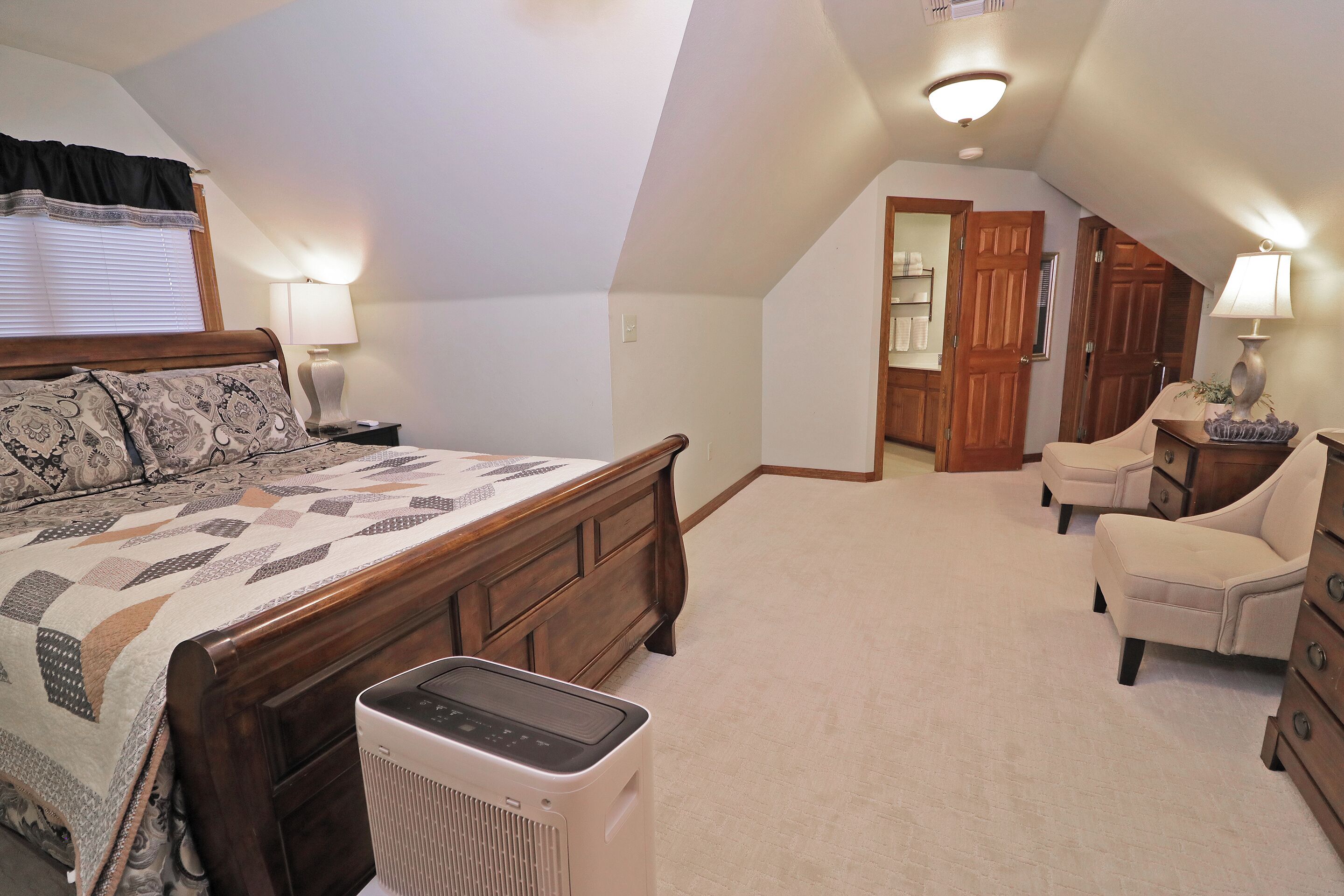 A bedroom with a bed, bedside lamps, a heater, a wooden dresser, a chair, and a door leading to another room.