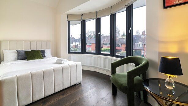 A modern bedroom with a curved window, a bed, a green armchair, and a table with a lamp. - Luxury 2BR Stay | Heart of London (London)