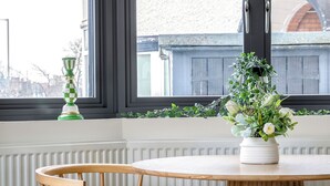 A wooden chair and table set against a window with a view of a building and a plant on the windowsill. - Luxury 2BR Stay | Heart of London (London)