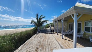A wooden deck with white chairs and a covered area with glass doors, overlooking a beach.
