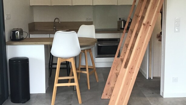 A modern kitchen with white cabinets, a wooden dining table with two chairs, a black trash bin, and a wooden ladder leaning against the wall.