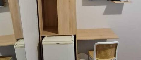 A compact room with a wooden desk, a white chair, a small white refrigerator, and a television mounted on the wall.