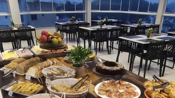 A buffet setup with various food items and utensils, arranged on a long table in a room with large windows and black chairs. - Hotel LF by Feliciano Joaçaba (Joaçaba)